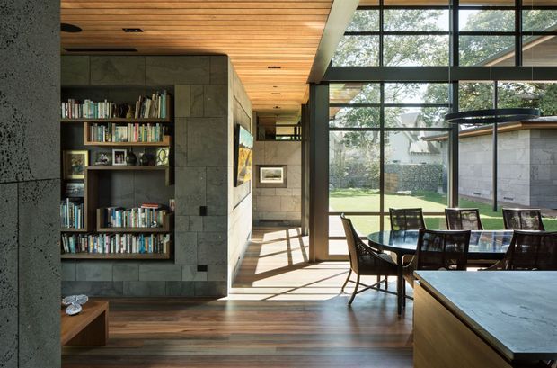 A bright contemporary open-plan dining and living area with a large timber dining table and dark chairs beside floor-to-ceiling glazing. A built-in dark stone or concrete shelving wall displays books, while the ceiling is clad in warm timber boards and the floor has dark timber flooring. The glazed wall opens views to a landscaped garden and provides strong natural light.