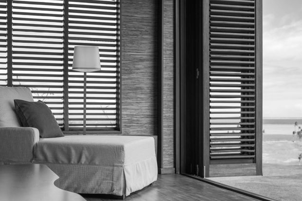 Black-and-white interior photograph of a calm lounge or sitting area beside the coast. A low upholstered chaise or sofa with cushions sits against full-height louvred windows, accompanied by a slim floor lamp. Textured wall cladding, dark timber flooring, and a large open louvred door frame an ocean view outside.