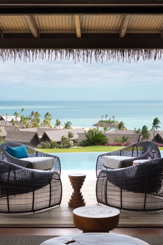 A tropical beachfront resort-style outdoor lounge viewed from beneath a thatched roof. Two dark woven armchairs with pale cushions face a turquoise swimming pool and expansive ocean horizon, with a low wooden table and stool between them. Additional thatched-roof buildings, palms, and landscaped gardens are visible beyond the pool. The scene is bright, relaxed, and coastal, with strong indoor-outdoor architectural framing.