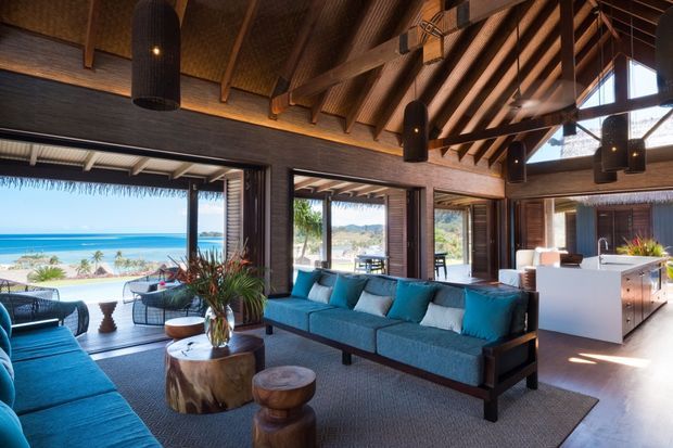 A spacious open-plan coastal living room with a dramatic exposed timber rafter ceiling, dark timber walls and framing, expansive glazed openings, and a large turquoise sectional sofa. The room opens directly onto a beachfront terrace with ocean views, while a white kitchen island is visible at the rear. Natural wood coffee tables, woven outdoor-style seating, and black pendant lights create a relaxed tropical-resort atmosphere.