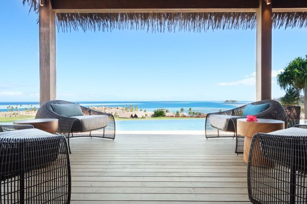 A bright open-air beachfront lounge beneath a substantial timber-framed pavilion with a thatched roof. The space has a broad pale timber deck, woven dark lounge chairs and sofas, small round timber tables, and unobstructed views across a sandy beach and blue ocean. Tropical planting and resort-style outdoor furnishings create a relaxed coastal setting.