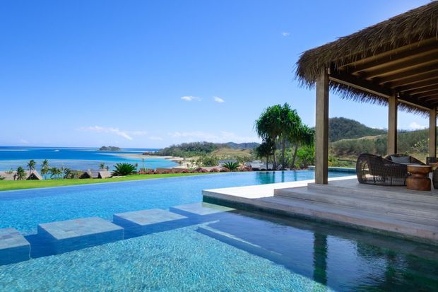 A tropical coastal outdoor living area with a large infinity-style swimming pool overlooking a bright blue ocean and small offshore islands. The pool includes submerged square platforms and steps, while a timber deck leads to a covered pavilion with sturdy timber posts, exposed rafters, and a thatched roof. Woven outdoor lounge furniture sits beneath the pavilion, with landscaped tropical planting surrounding the pool.