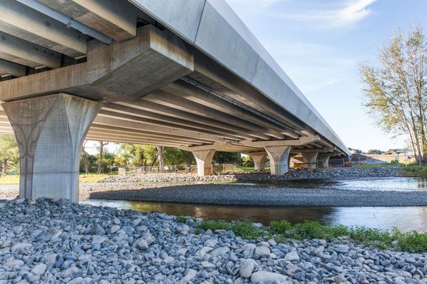 A low-angle exterior view beneath a large concrete bridge spanning a shallow rocky waterway. Repeated sculptural concrete piers support the broad deck, with exposed structural beams and a metallic edge fascia visible along the bridge. The surrounding landscape includes gravel banks, shallow water, trees, and a distant road infrastructure.