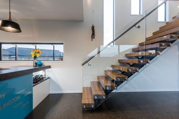 Bright contemporary open-plan interior showing a modern kitchen edge and a floating timber staircase with glass balustrades and dark metal supports. The staircase rises beside tall windows and a high void, with polished dark flooring, white walls, and a black pendant light. A partial kitchen island with dark stone benchtop and turquoise cabinetry is visible at left.