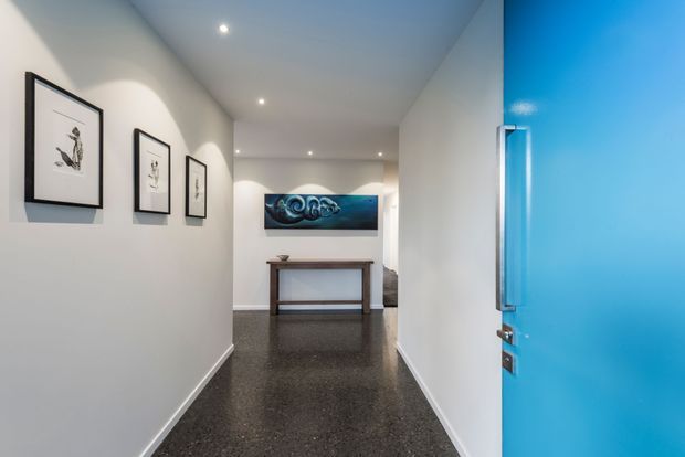 A modern residential hallway viewed from an open bright blue entry door. The corridor has smooth white walls, recessed ceiling downlights, dark polished flooring, framed artwork, and a narrow timber console table beneath a blue abstract artwork.