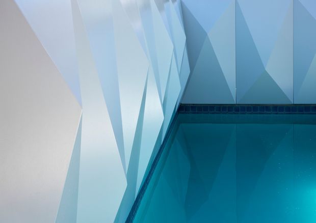 Close-up architectural detail of a swimming pool corner, showing vivid blue water, a dark tiled pool edge, and sharply faceted geometric wall or ceiling surfaces in pale blue and white. The abstract angular forms create a contemporary, sculptural atmosphere.