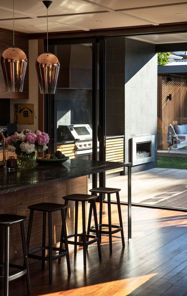 A stylish open-plan kitchen and entertaining area opening onto a landscaped outdoor courtyard. The kitchen features dark timber cabinetry, a glossy dark stone island bench with bar seating, black-framed glazed doors, and two reflective copper pendant lights. Beyond the fully opened doors is an outdoor barbecue, tiled fireplace wall, timber privacy screening, outdoor seating, and decking. Strong sunlight creates dramatic shadows across the timber floor and patio.