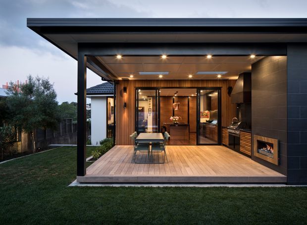 A contemporary covered outdoor entertaining pavilion opening onto a landscaped lawn. The structure has a broad flat roof with recessed downlights, dark exterior cladding and framing, timber-lined soffits and decking, a glazed connection to the interior, an outdoor dining setting, built-in barbecue, and a modern fireplace integrated into the side wall.