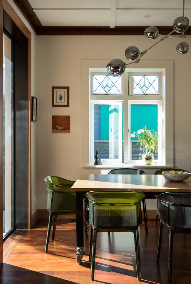A refined dining area with a rectangular timber dining table and translucent olive-green dining chairs, positioned beside a tall multi-pane window. The room features polished dark timber flooring, cream walls, white coffered ceiling detailing, dark timber ceiling beams and trims, framed artwork, and a sculptural chrome multi-arm pendant light. Bright turquoise exterior cladding is visible through the window.