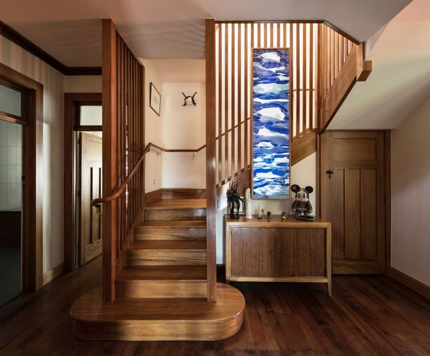 Warm, wood-lined residential stair hall with a curved-bottom staircase, vertical timber slat balustrade, dark timber flooring and trim, and a mid-century-style timber console beneath a tall blue-and-white artwork. A hallway and timber-panelled door are visible alongside the stair.