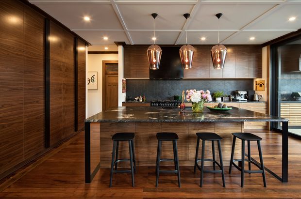 A sophisticated contemporary kitchen with dark timber wall panelling and cabinetry, a large stone island with seating for four, and a dark textured splashback. Three pendant lights hang above the island, while warm recessed lighting and a coffered ceiling create a polished, inviting atmosphere. Timber flooring and matching bar stools reinforce the warm, refined material palette.