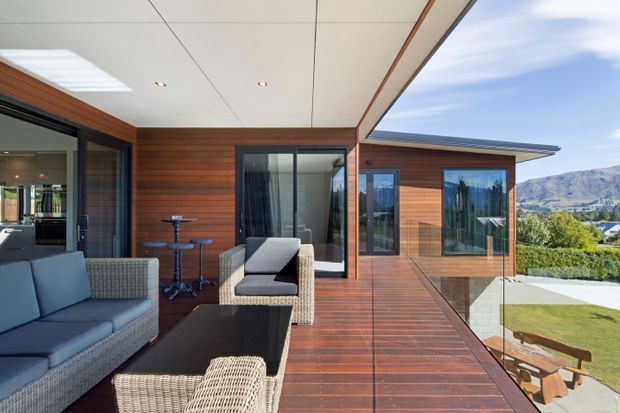 A contemporary timber-clad house with a covered outdoor terrace and balcony-style deck. The space features wicker lounge furniture, a dark outdoor coffee table, large glazed sliding doors, timber decking, and frameless glass balustrades. The terrace overlooks a landscaped lawn and mountainous terrain, with warm timber surfaces contrasting against cool grey upholstery and a clear blue sky.