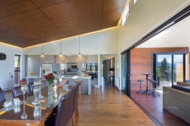 Open-plan contemporary interior featuring a dining area and kitchen with extensive timber finishes. A large dining table with upholstered chairs occupies the foreground, while a kitchen island, stainless-steel appliances and pendant lights sit beyond. The space opens through wide glazed sliding doors to a timber-lined outdoor entertaining area, with warm timber flooring and a timber ceiling creating visual continuity.