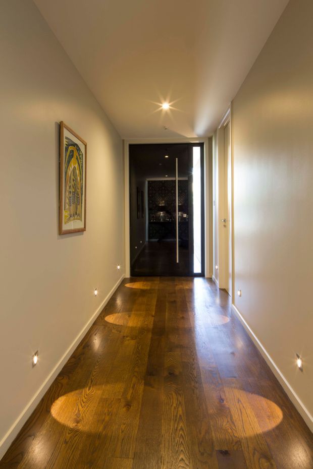 A long, narrow residential hallway with warm white walls, a polished dark timber floor, recessed low-level wall lights, and a framed artwork. Several white doors open off the corridor, leading toward a dark glazed doorway at the far end. The space has a clean, contemporary feel with warm, focused lighting.