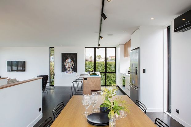 A bright, open-plan contemporary kitchen and dining area. The foreground features a large light timber dining table with black chairs, glassware and a simple floral arrangement. Behind it is a compact kitchen with a timber island, white cabinetry, integrated appliances, dark flooring, black-framed glazing and track lighting. A living area is visible to the left, with artwork and a television.