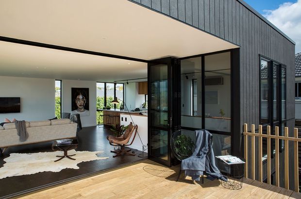 A contemporary residential extension viewed from a timber deck, with a dark vertical-clad glazed pavilion opening into a bright open-plan living and kitchen area. The interior includes mid-century-style lounge seating, a pale sofa, timber cabinetry, polished dark flooring and extensive glazing. A timber balustrade frames the deck, while the projecting roof creates a sheltered indoor-outdoor connection.