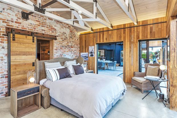 A spacious rustic-industrial bedroom with a large upholstered bed, exposed white-painted timber roof trusses, a pale timber ceiling, warm timber wall panelling, and a feature wall of exposed brick. Sliding barn-style timber doors, woven bedside and lounge furniture, polished concrete flooring, and extensive glazed openings create a relaxed warehouse-conversion feel.