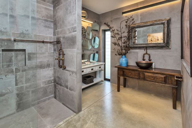A spacious industrial-style bathroom featuring a large walk-in tiled shower with exposed plumbing, a floating vanity with twin rounded mirrors, and a dark timber console supporting a vessel basin. Grey concrete-look wall and floor finishes, aged metal fixtures, warm concealed lighting, and a large framed mirror create a refined, moody atmosphere.