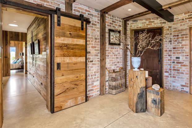 Rustic industrial-style entry hall featuring exposed red brick walls, warm timber ceiling beams and wall cladding, a large sliding barn door with black metal hardware, and a dark timber entry door. Decorative natural wood stump pedestals, a woven basket, and branches add organic character. A hallway and glimpse of a bedroom extend beyond the open doorway.