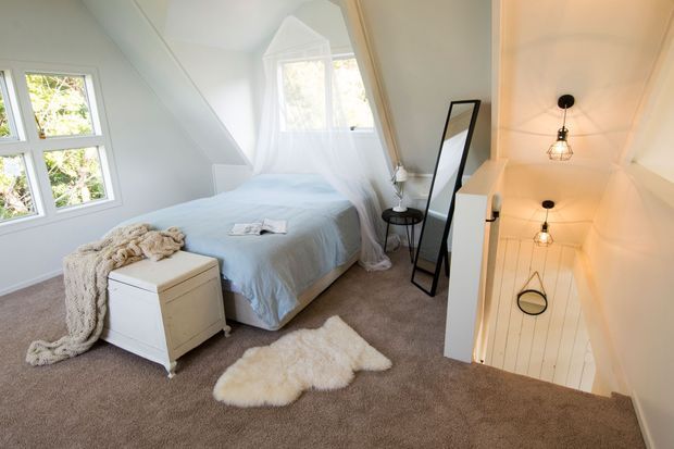 Bright attic-style bedroom with a low sloping ceiling, white-painted walls and timber details, soft blue bedding, a white chest, neutral carpet, and a small stairwell opening with pendant lights. A large mirror and windows bring additional light into the compact space.