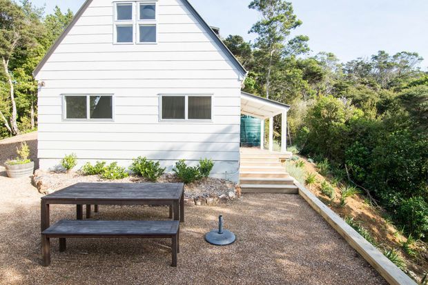 Exterior view of a compact white weatherboard gable-roofed house set within dense bush landscaping. The facade has horizontal cladding, dark-framed windows, and a side entrance reached by timber steps, with a gravel outdoor area and picnic-style table in the foreground.