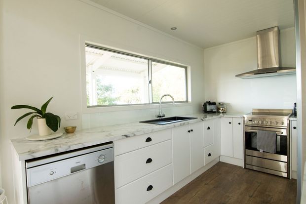 Bright white galley-style kitchen with shaker-style cabinetry, marble-look benchtops, stainless-steel appliances, a farmhouse-style sink, timber-look flooring, and a large horizontal window above the sink. The space has a clean, classic coastal feel with natural light and restrained detailing.