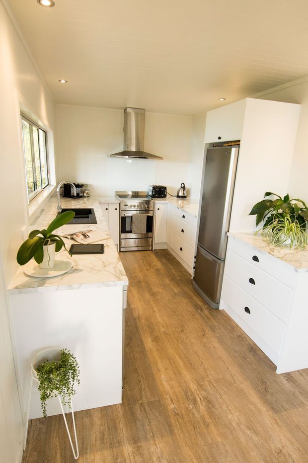 Bright, compact galley-style kitchen with white cabinetry, a marble-look benchtop, stainless-steel appliances, timber-look flooring, recessed ceiling lights, and a large window providing natural light. Potted plants add greenery, while the clean white-and-wood palette creates a fresh contemporary feel.