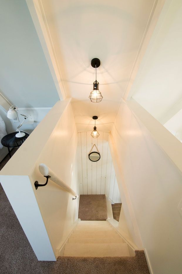 A narrow residential stairway descending from an upper landing, with cream carpeted stairs, white painted walls and ceiling, vertical white wall panelling, dark metal handrails, and a series of warm pendant lights. A small adjacent living area is partially visible at the upper left.