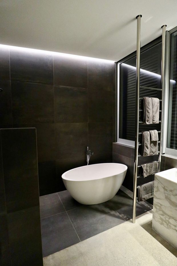 A contemporary dark-toned bathroom featuring a freestanding white oval bathtub against large charcoal wall and floor tiles. A chrome heated towel rail stands beside the bathtub, with a marble-look vanity partially visible on the right. Integrated linear lighting along the ceiling edge and black-framed glazing create a refined, moody atmosphere.