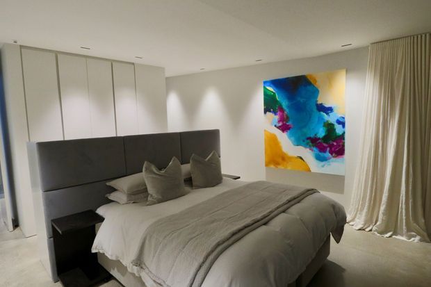 A contemporary bedroom with a large upholstered charcoal-grey bedhead, layered neutral bedding, matching dark bedside table, floor-to-ceiling cream curtains, built-in white wardrobes, recessed ceiling lighting, and a vibrant abstract artwork on the wall. The room has a soft, refined neutral palette with colourful art as the main accent.