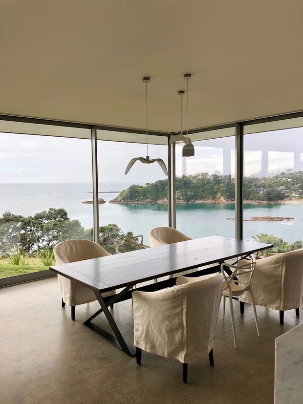 A contemporary dining area with a long dark timber dining table and upholstered dining chairs, set against expansive floor-to-ceiling corner glazing. The room overlooks a calm coastal inlet with rocky shoreline and lush vegetation. Minimal pendant lights and a sculptural suspended bird-like feature complement the warm, understated interior.