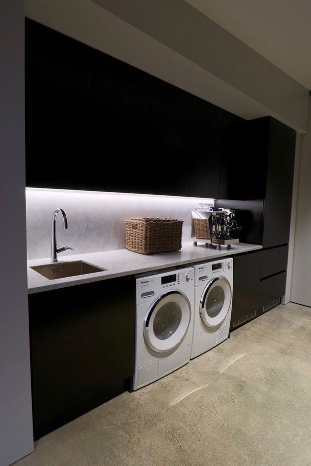 A sleek contemporary laundry room with matte black full-height cabinetry, a pale stone benchtop and integrated backsplash illuminated by under-cabinet lighting. The room includes a stainless-steel sink with curved tap, two front-loading white appliances, woven storage baskets, and a coffee machine. The floor is finished in polished concrete, creating a dark, refined and functional interior.
