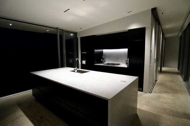 A sleek contemporary kitchen dominated by a large white stone island with integrated sink and tap. Gloss-black cabinetry and a dark splashback surround the cooktop and illuminated recessed rangehood, while full-height glazing and a concrete-look floor extend toward a hallway. Recessed ceiling lighting and minimal detailing create a refined, high-contrast interior.