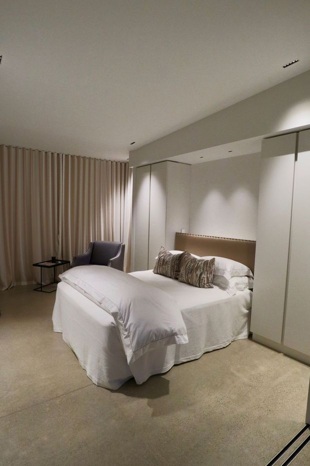 A softly lit, contemporary bedroom with a neatly made bed, upholstered beige headboard, patterned accent cushions, full-height wardrobes, floor-to-ceiling beige curtains, a dark lounge chair, and a small side table. The room has a calm neutral palette and a carpeted floor.