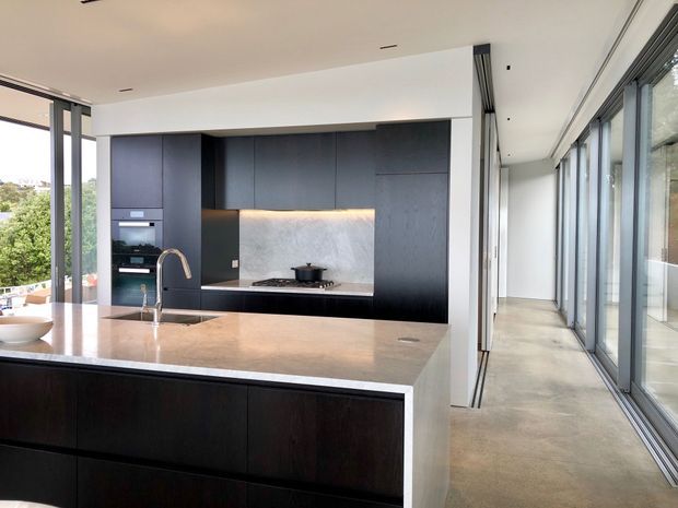 A sleek contemporary kitchen with a large stone island, dark timber-look cabinetry, integrated appliances, a pale stone splashback and broad floor-to-ceiling glazing. The kitchen opens onto a long, bright glazed corridor or living edge with views to greenery outside.