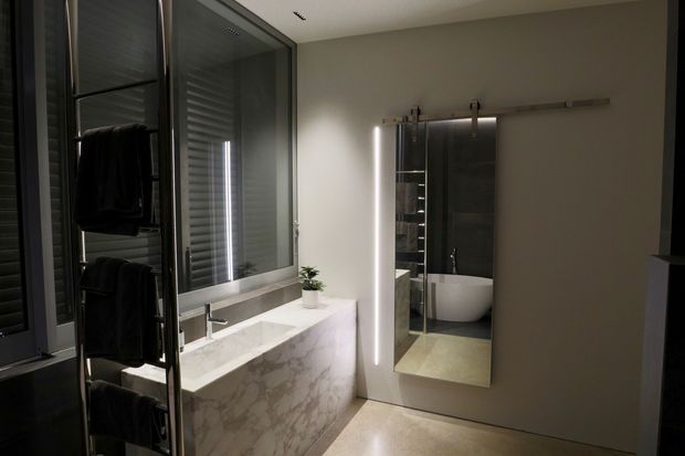 A contemporary bathroom with a sculptural freestanding stone-clad vanity, integrated basin and chrome tapware. A large illuminated sliding mirror reflects a freestanding white bathtub and dark tiled shower area. Full-height glazing with horizontal blinds and a heated towel rail contribute to the refined, minimalist fit-out.