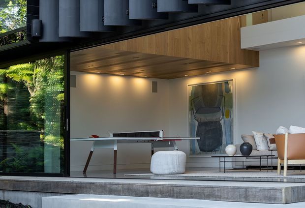 A refined contemporary living room opening onto a lush garden through expansive black-framed glazing. The room features a warm timber-lined ceiling and overhead built-in cabinetry, white walls, a minimalist glass dining or coffee table, sculptural seating, a low black metal console, and a pale upholstered sofa with tan leather accent chairs. Layered recessed lighting creates a calm indoor-outdoor atmosphere.