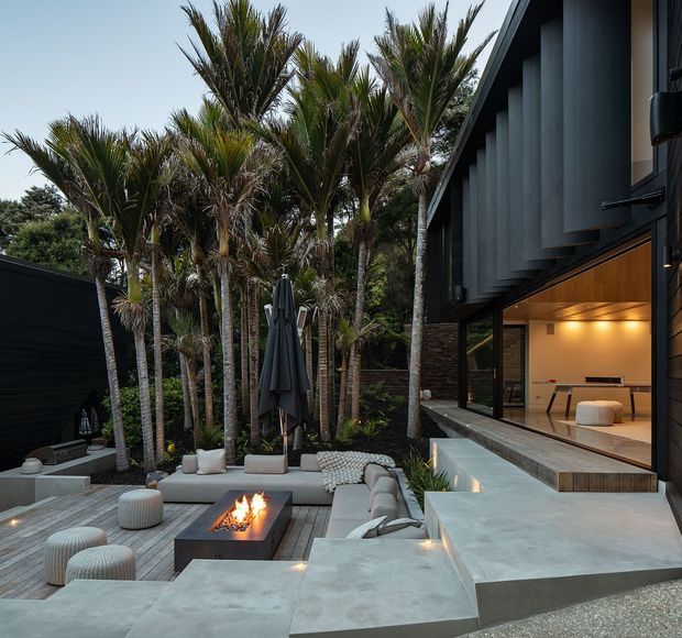A contemporary multi-level outdoor entertaining area beside a dark-clad modern house, featuring a sunken lounge with a rectangular fire table, pale upholstered seating, timber decking, concrete steps and platforms, and dense tropical planting. Large glazed openings reveal a warmly lit interior with minimalist cabinetry and furnishings. The exterior composition includes strong vertical dark cladding and an elevated indoor-outdoor connection.