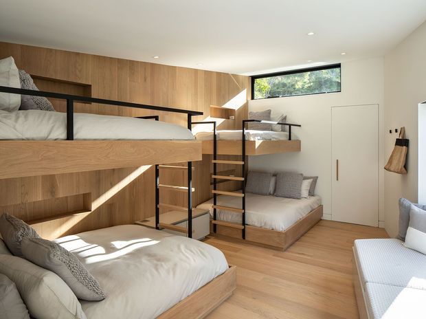 Bright compact bedroom with custom timber bunk-style sleeping platforms, integrated storage, black metal guardrails and ladder, a built-in lower sofa/daybed, and light timber flooring and wall panelling. Large windows and skylights provide natural light, creating a warm, efficient multi-sleeping space.