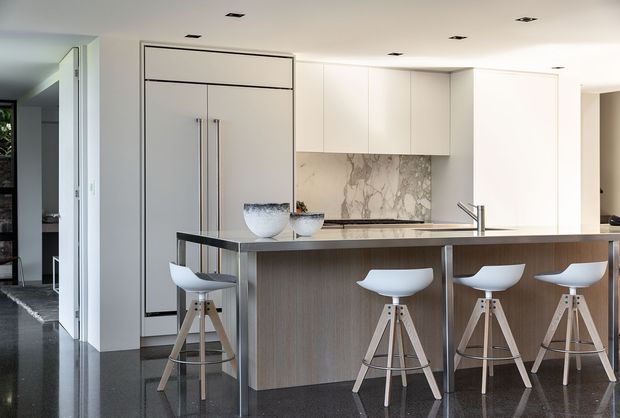 Bright contemporary kitchen with a large island and four sculptural bar stools. The space features white full-height cabinetry, integrated refrigerator doors, a marble-look splashback, dark polished flooring, and a timber-look island front with a slim dark benchtop. Recessed ceiling downlights and concealed appliances create a clean, refined appearance.