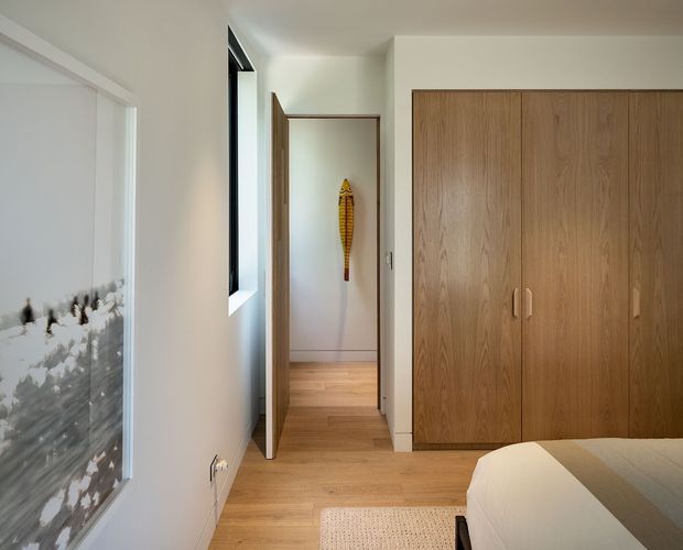 Bright, minimalist bedroom interior with pale timber flooring, a large built-in natural wood wardrobe, white walls, and a partially visible bed. A doorway leads to a small hall or adjoining space, while artwork and a narrow window add visual interest.