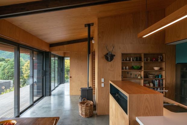 A warm, wood-lined open-plan kitchen and circulation space with extensive floor-to-ceiling glazing along one side. The kitchen features a timber island with integrated dark feature panels, open shelving, a concealed or integrated appliance bank, and a compact black wood-burning stove near the glazed corridor. Polished concrete flooring and exposed dark ceiling beams create a refined contemporary cabin-like interior with strong indoor-outdoor visual connection.