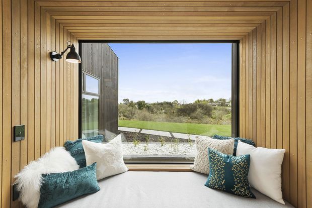 Compact window seat or reading nook enclosed in warm vertical timber panelling, with a deep cushioned built-in bench, layered white and teal cushions, and a large picture window overlooking a green landscape. The ceiling and walls feature detailed concentric timber trim around the opening, with a small wall-mounted reading light.