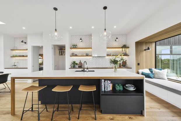 Bright contemporary kitchen with a large white island featuring a dark integrated base, timber breakfast bar and open shelving. The room includes white cabinetry, a white patterned tile splashback, timber flooring, pendant globe lights, black tapware and stools. A built-in window seat and large glazed opening are visible on the right, with a living area beyond.