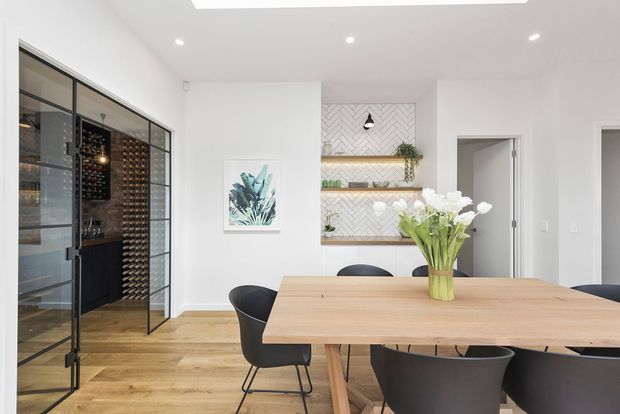 Bright contemporary dining area with a large light timber dining table and black upholstered chairs. A glazed, black-framed wine storage room is visible on the left, while the rear wall features open timber shelving, a white herringbone tile backing, and integrated display lighting. White walls, light timber flooring, recessed ceiling lights, and a skylight create an airy, refined interior.