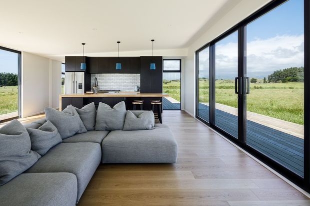 Contemporary open-plan living room with a large grey sectional sofa facing a compact kitchen. The space has pale timber flooring, white walls and ceiling, dark built-in kitchen cabinetry, a light tiled splashback, pendant lights, and extensive black-framed floor-to-ceiling glazing opening onto a timber deck and grassy landscape.