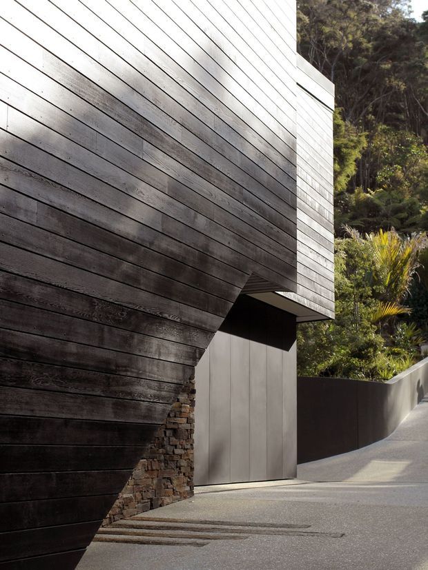 Close-up exterior architectural view of a contemporary, cantilevered building clad in dark horizontal timber boards. The projecting upper volume is supported above a recessed entry or garage-like opening, with a rough stacked-stone base and dark metal wall panels below. A sloping paved driveway or walkway leads past landscaped native vegetation and dense trees.