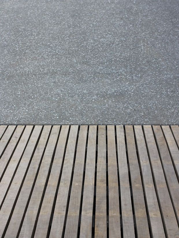 Close-up view of a weathered timber deck or boardwalk meeting a grey textured paved or concrete surface. The image is abstract and material-focused, with parallel wooden decking boards in the foreground and a contrasting rough grey surface beyond.