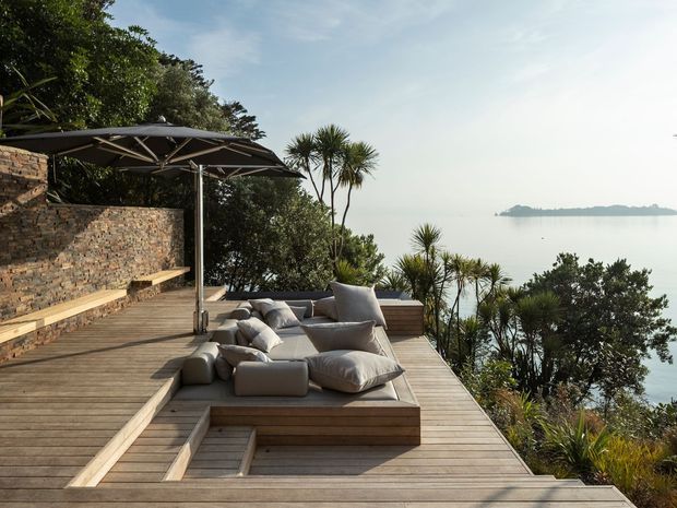 A timber-clad waterfront terrace stepping down toward calm water, with built-in low timber seating, loose outdoor cushions, a large cantilevered shade umbrella, and a textured stacked-stone retaining wall. Native-looking planting borders the deck, and a distant island or shoreline is visible across the water. The setting feels relaxed, natural, and contemporary.