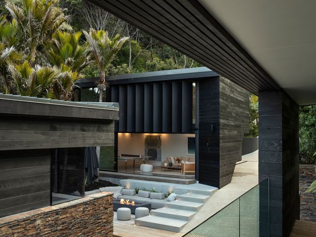 A modern multi-level residence nestled in dense tropical vegetation. The view looks across dark horizontal timber-clad volumes, a projecting flat roof, vertical dark facade fins, and a stone-clad wall toward a glazed living area. Broad concrete steps lead down to a sunken outdoor seating and fire feature, with a glass balustrade along the elevated walkway.